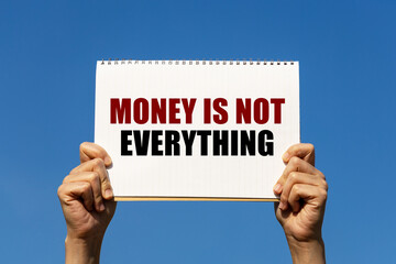 Money is not everything text on notebook paper held by 2 hands with isolated blue sky background....