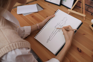 Fashion designer creating new look. Woman drawing sketch in book with pencil at wooden table,...