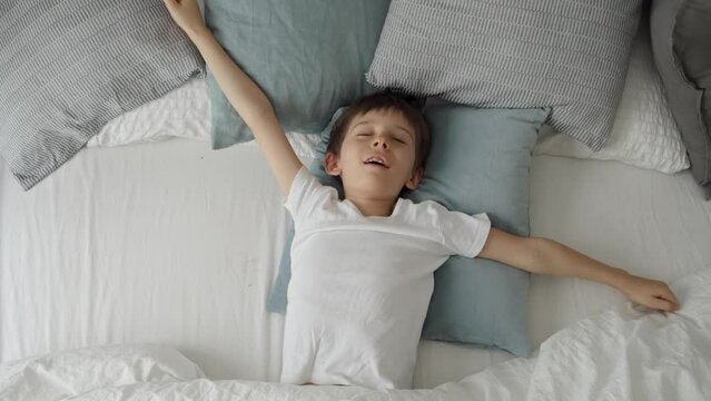 Cute little boy lying in bed, stretching hands and yawn. Child waking up in bed at morning
