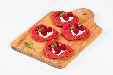Viennese Red heart-shaped waffles with cream cheese and raspberries, decorated with micro greenery. Waffles with the addition of beet juice. On a wooden board. White background
