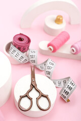 Podiums with scissors, thread spools and measuring tapes on pink background, closeup