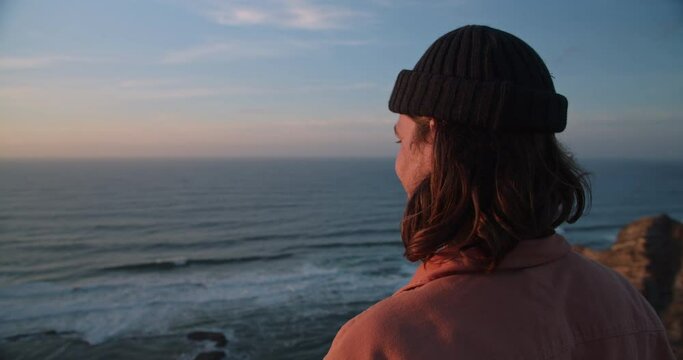 Young, bearded man watching the sunset and waves rolling in along the coastline and cliffs of the Western Algarve, Portugal