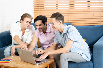 Triple young man sitting in the living room while using laptop to video call with rainbow flag on desk. LGBTQ people lifestyle and love emotion. LGBT social network. selective focus