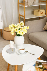 Vase with flowers and cup of coffee on table in living room