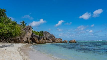 Fototapeta na wymiar Picturesque granite boulders and rocks with steep smoothed slopes on a tropical beach. Turquoise ocean waves on the sand. Lush tropical vegetation. Blue sky, clouds. Seychelles. La Digue. 