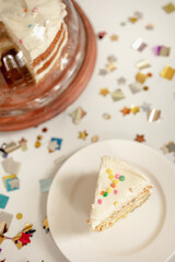 Obraz na płótnie Canvas Confetti or Funfetti Cake Slice with Colorful Fun Sprinkles and Three Layered Cake in Covered Cake Stand in the Background surrounded by Metallic Confetti Shapes 