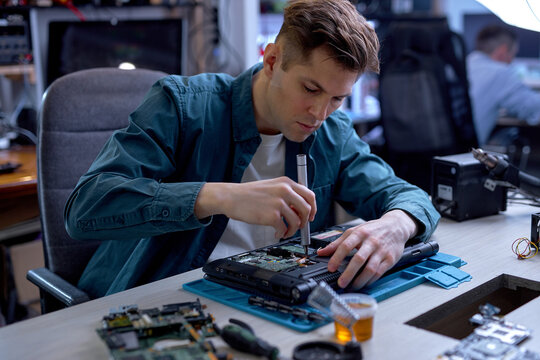 confident guy repairing parts in disassembled broken laptop using screwdriver and different tools on table in workshop, professional technician at work place, concentrated on repair
