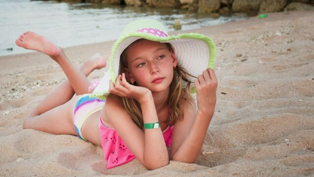 Pensive smiling lovely blonde teen girl relaxing enjoying lying on sandy beach near ocean. Tourist teenager in sunhat and swimsuit tanning and resting on resort. Vacation, travel, tourism concept.