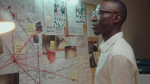 Professional black detective standing in his office at night, examining photos, map and notes connected by red thread on investigation board, thinking about crime