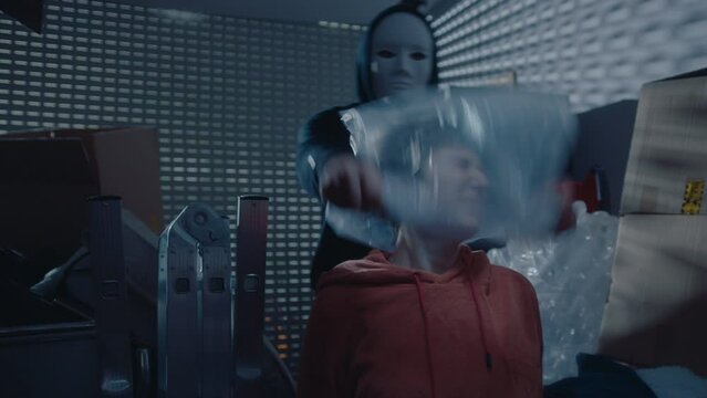 Violent murderer in mask and black hoodie choking struggling female victim tied to chair with plastic bag on her head in dark garage at night. Cinematic medium shot