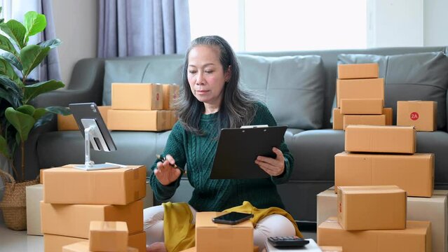 Senior Asian business owner woman prepare parcel box and standing check online orders for deliver to customer on tablet, laptop Shopping Online concept..