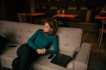 Tired blonde businesswoman relaxing on a sofa at the coffee bar after working remotely with her colleagues all day. She is listening to them having a conversation