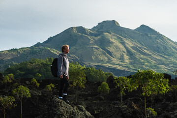 Fototapeta premium A young girl traveler with a backpack walks in the mountains against the backdrop of the Batur volcano on the island of Bali in Indonesia.