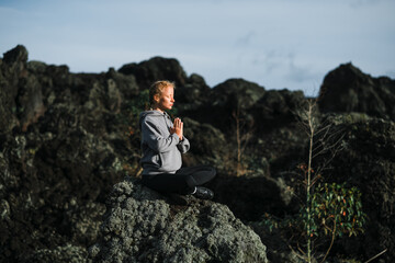 Naklejka na ściany i meble A young beautiful girl, a yogini, sits in the mountains, with her eyes closed, clasped her hands in namaste and meditates.