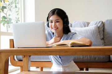 Portrait of a student learning on line with headphones and laptop taking notes in a notebook sitting at her desk at home.