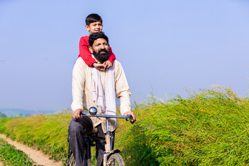 Rural Indian father and son riding bicycle in agricultural field in bright sunny day, Parents with...