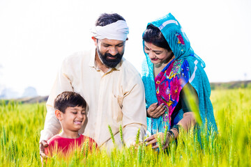 Portrait of happy young indian farmer family touching wheat crop standing at agriculture field in bright sunny day. Parent with child ,Rural india concept.