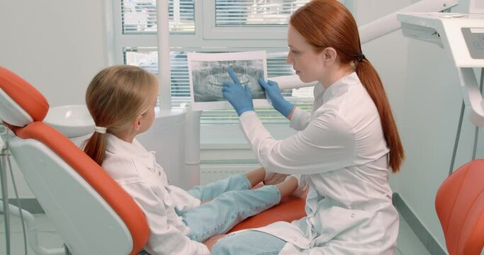 professional talented female doctor with a dental picture. show a x ray picture of a teeth to little pretty blonde girl.