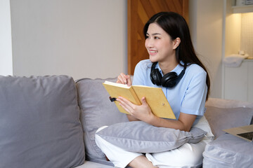 Bright and cute Asian woman sitting on sofa with laptop to work and happily taking notes at home.