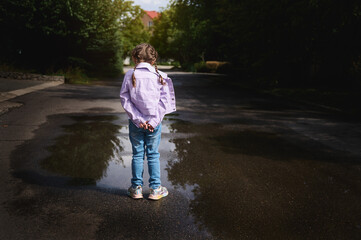 Little girl in jeans and purple jacket, standing with her hands crossed by a small puddle after rain and looking at her reflection in the water while walking on the countryside street. Rear view