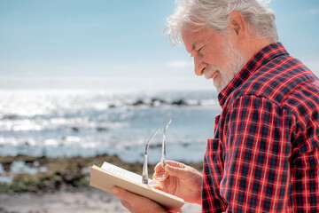 Portrait of senior bearded man reading a book in front to the sea. Elderly relaxed pensioner with eyeglasses enjoying retirement in outdoor