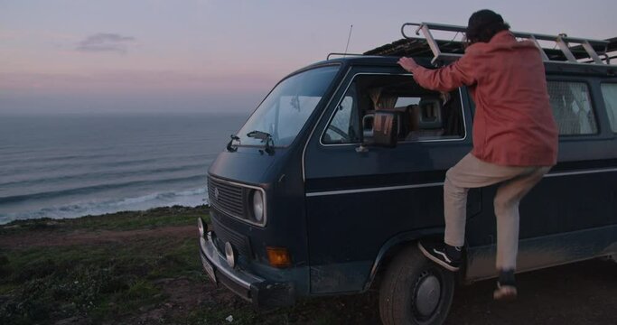 Young man watching and celebrating the sunset on the roof of his vintage van during golden hour along the cliffs and coastline of the Western Algarve, Portugal