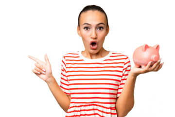 Young Arab woman holding a piggybank over isolated background surprised and pointing side