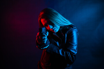 A young beautiful woman with long hair in a black leather jacket is holding a gun in the dark at...