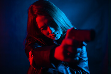 A young beautiful woman with long hair in a black leather jacket is holding a gun in the dark at...