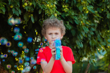 A curly-haired boy in a red T-shirt plays with a gun that generates soap bubbles. Children's game in summer. Educational toys