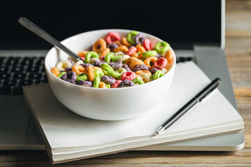 Colored breakfast cereals in a bowl and laptop, workplace breakfast, quick healthy breakfast in the office in the morning.