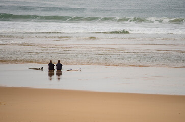 Couple of surfers sitting on the sand facing the Atlantic Ocean
