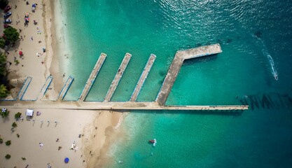 Aerial view of a pier extending out over crystal blue waters and people sunbathing on the shore