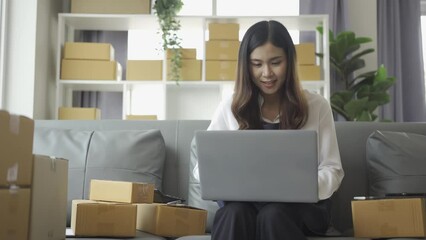 Female entrepreneur working at home office with smartphone with laptop, tablet, deck (SME business). Business concept of Asian woman checking customer orders at home, delivering goods to customers.
