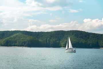 Naklejka premium Tranquil scene with a sailing boat on a calm lake with forest hills in the background. Recreational boating on the lake in summer
