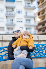 Young mother with her cute infant baby boy child on bench in city park