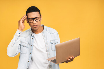 Young serious thinking tired stressed african american man standing and using laptop computer isolated over yellow background.