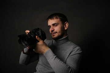 Portrait of a young handsome man in a gray jacket with a camera in his hand. The photographer on a black background sits on a chair.