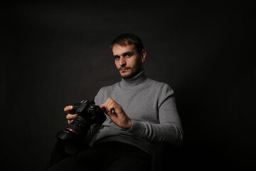 Portrait of a young handsome man in a gray jacket with a camera in his hand. The photographer on a black background sits on a chair.