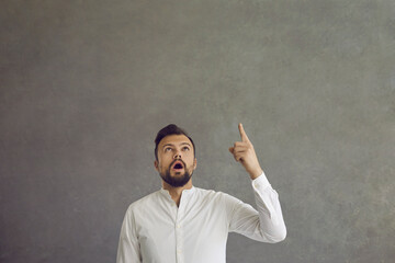 Young man with a surprised expression on his face is stunned by a thought or idea that came to his mind. Man looks up and points his finger at the free space for text on a gray background. Banner.