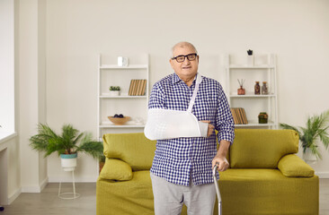 Portrait of a senior gray-haired elderly man standing at home in rehabilitation with crutch and with arm in a plaster cast after injury. Patient wearing sling or bandage. Recovery and rehab concept.