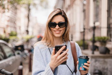 Beautiful smiling woman walking on street with coffee takeaway, chatting on mobile phone. Girl...
