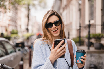 Fototapeta premium Beautiful smiling woman walking on street with coffee takeaway, chatting on mobile phone. Girl talking on smartphone on her way to meeting spot, sending message, wear blue sweater and black glasses. 