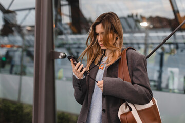 Attractive blonde woman standing on bus station and wait for a bus, using mobile phone, read sms,...