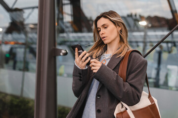 Attractive blonde woman standing on bus station and wait for a bus, using mobile phone, read sms, text, news. Portrait of calm confident female blogger using cellphone reading.