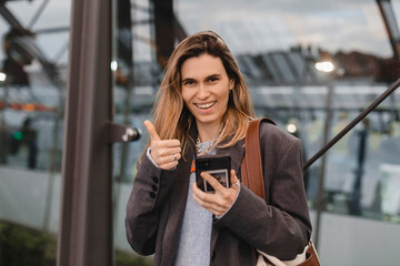 Portrait of a pretty casual girl holding mobile phone and showing thumbs up standing on bus...
