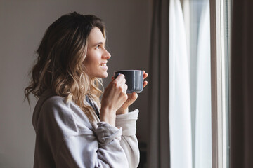 Close-up portrait of laughing blonde girl in beige hoody drinking coffee standing near window. Lovable lady standing near window and looking at beautiful building with cup of tea or coffee.