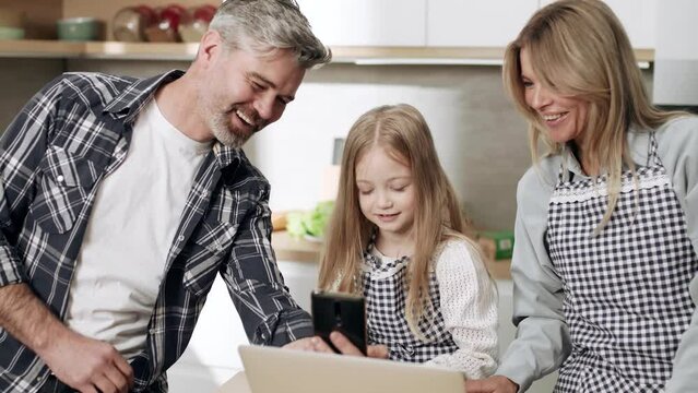 Smiling mature man show funny picture or video on phone to wife and daughter while they browsing studying on laptop computer indoors Happy family laughing and spend time together at domestic kitchen
