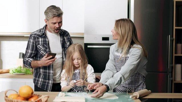 Smiling mature man approaches his charming wife and cute little daughter while they prepare cookies for baking
and takes selfie on smartphone Happy family laughing and spend time together at kitchen