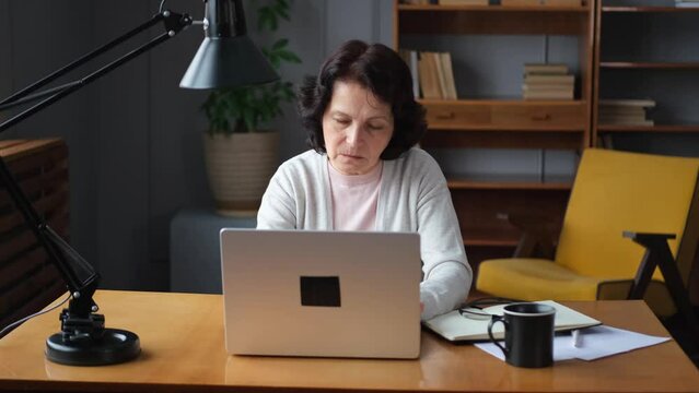 Confident stylish european middle aged senior woman using laptop at home. Stylish older mature 60s lady sitting at table looking at computer screen typing chatting reading writing email